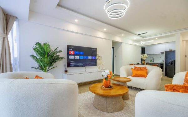 Modern living room with neutral furniture, orange accents, circular coffee tables, and a flat-screen TV on the wall.