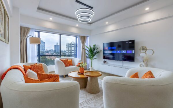 Modern living room with white curved sofas, orange cushions, a round wooden coffee table, large windows, and a wall-mounted TV.