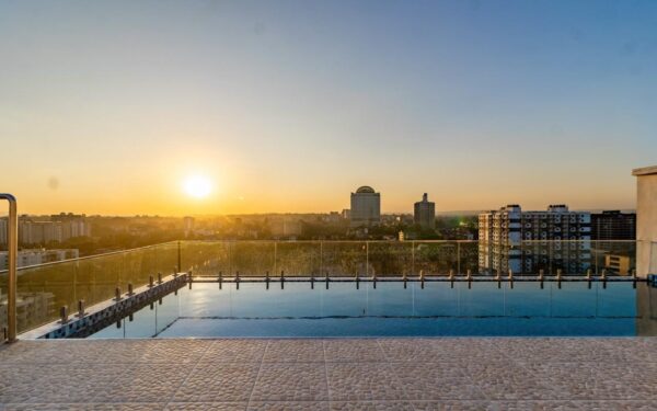 Sunset over city skyline viewed from a rooftop pool with glass barrier and textured stone tiles in the foreground.