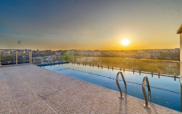 Rooftop infinity pool overlooking a cityscape at sunset, with a clear sky and railings in the foreground.