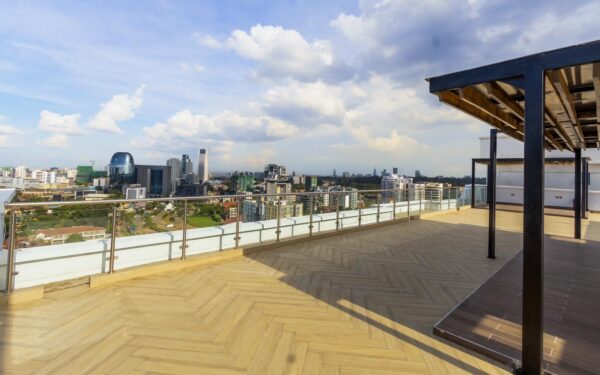 Rooftop terrace with wooden flooring overlooks a city skyline under a partly cloudy sky. Modern buildings are visible in the distance.