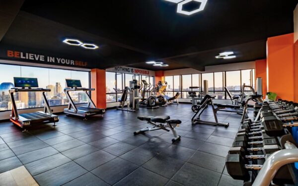 Spacious gym with treadmills, weight machines, and dumbbells, large windows showing city skyline, motivational quotes on walls.