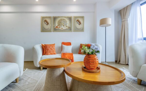 Modern living room with white sofa, orange pillows, abstract art, wooden tables, and a vase with flowers near a tall lamp.