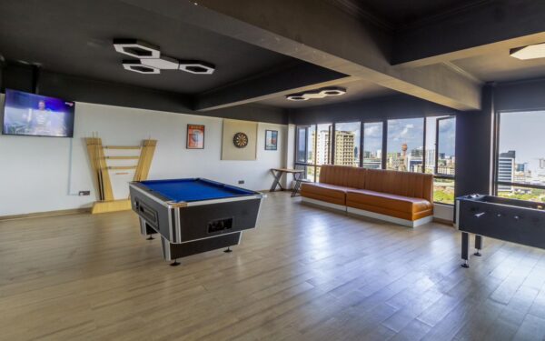 Game room with blue pool table, dartboard, foosball table, brown sofa, flat-screen TV, and windows showing city skyline.