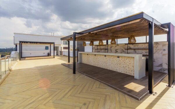 Spacious rooftop terrace featuring a modern pergola with a tiled bar counter, wooden deck, and glass railing under cloudy skies.