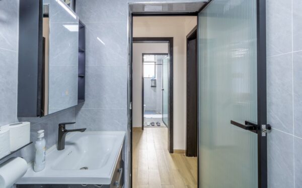 Modern bathroom with gray tiles, white sink, and black faucet. Open door reveals a shower and window with natural light.