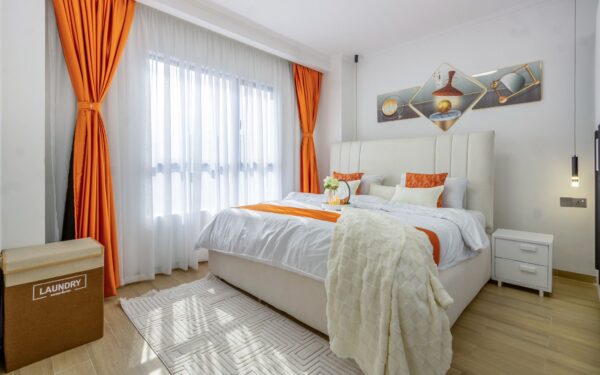 Bright bedroom with orange accents, white bed, geometric wall art, orange curtains, and a laundry bin by a large window.
