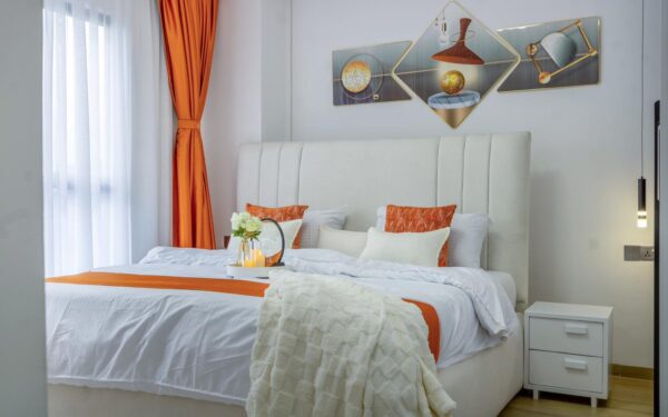 Cozy bedroom with white bed, orange accents, tray with flowers, modern art on wall, orange curtains, and pendant light.