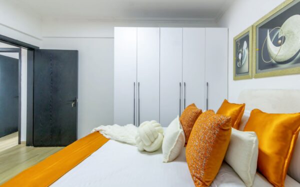 Bedroom with white wardrobes, a bed decorated with orange and white pillows, an orange throw, and abstract wall art above the headboard.